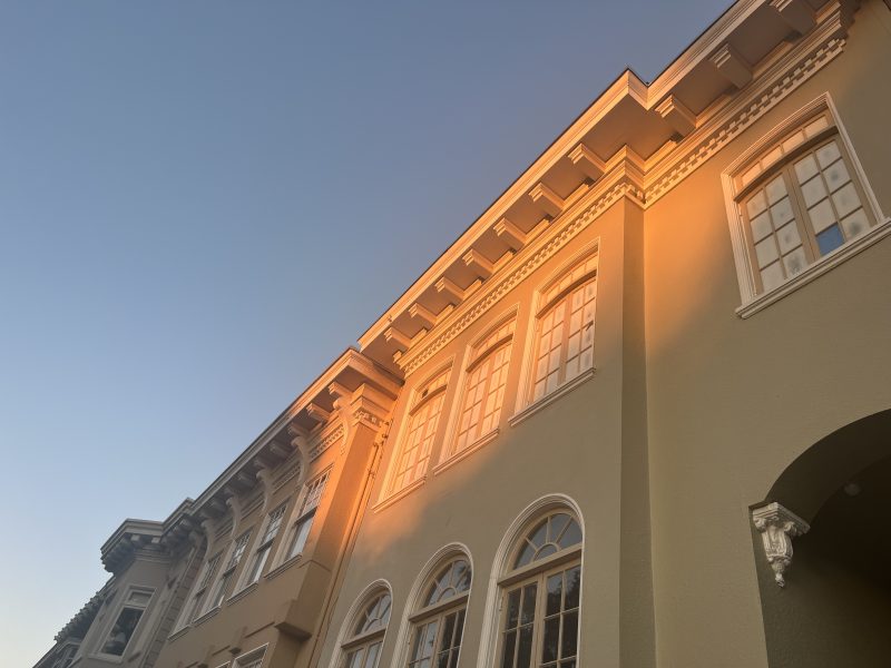 A beige building with large windows is illuminated by warm sunlight, set against a clear blue sky.