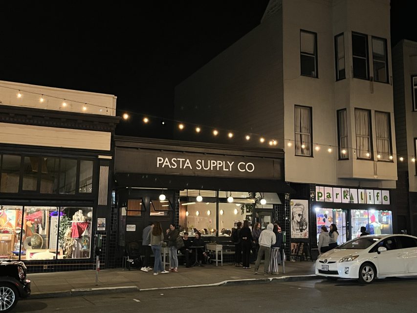 Nighttime view of Pasta Supply Co restaurant with people dining outside; string lights are hung above and parked cars line the street.