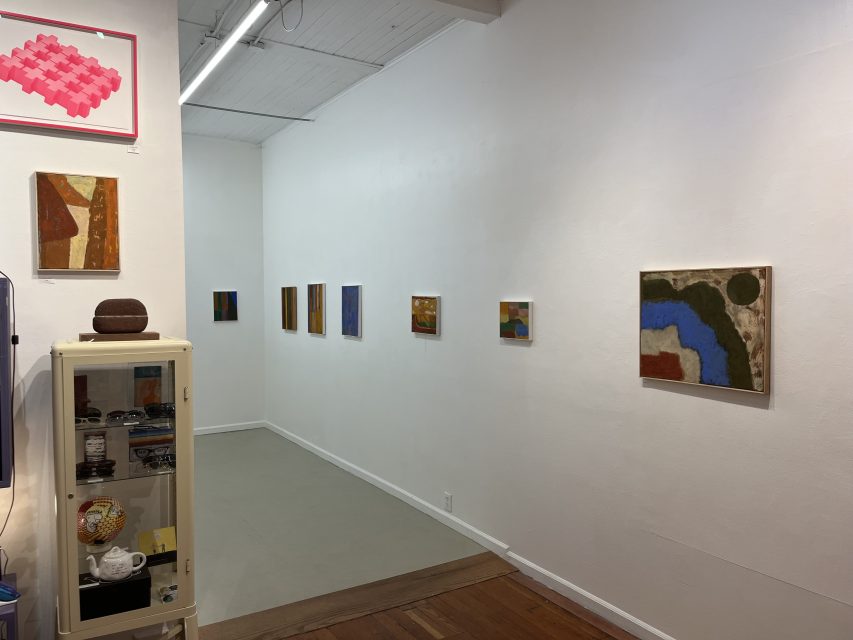 A small art gallery room with abstract paintings on white walls and a display cabinet containing small items in the corner.