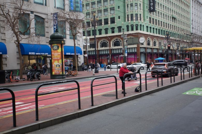 A city street with people walking on the sidewalk, a cyclist in a red jacket riding along a bus lane, and shops in the background, including a Ross Dress for Less store.
