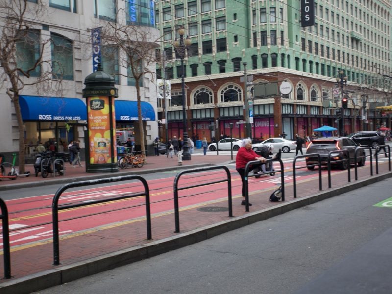 A city street with red bus lanes, pedestrians on the sidewalk, and storefronts including Ross Dress for Less. A person in a red jacket sits on a railing in the foreground.