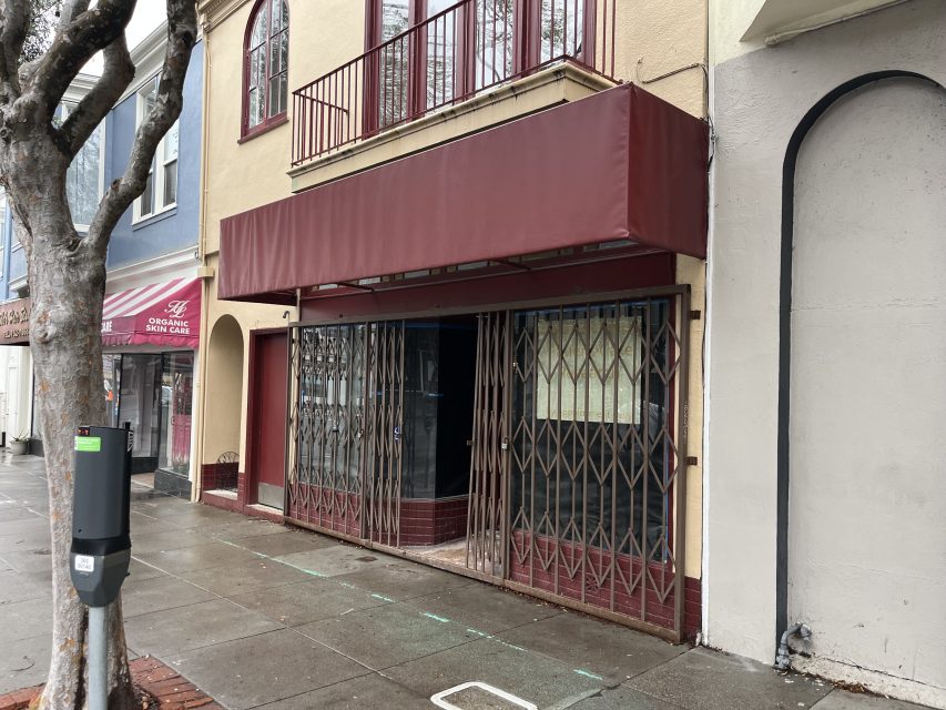 A storefront with a red awning and metal security gates is closed on a wet sidewalk alongside other businesses.