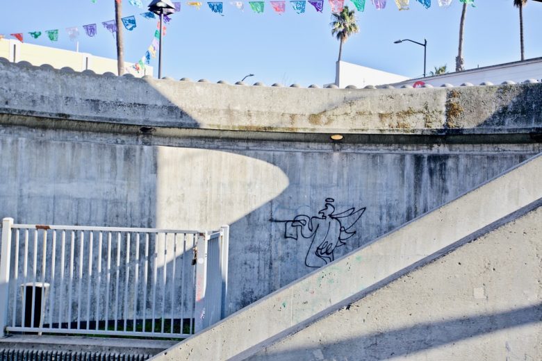 Graffiti of an angel with a trumpet is drawn on a curved concrete wall, with colorful flags strung above and shadows cast on the structure.