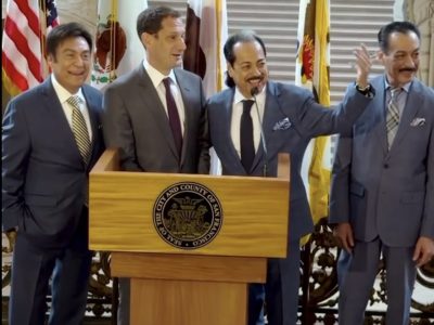 Four men in suits stand behind a podium with the Seal of San Francisco, posing for a photo with flags and window blinds in the background.