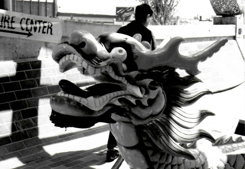 A close-up of a dragon sculpture outdoors, with a person and a building in the background. The image is in black and white.