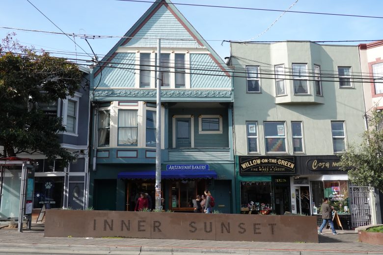 Street view of shops and apartments in the Inner Sunset neighborhood, including Arizmendi Bakery and Willow on the Green, with pedestrians walking by.