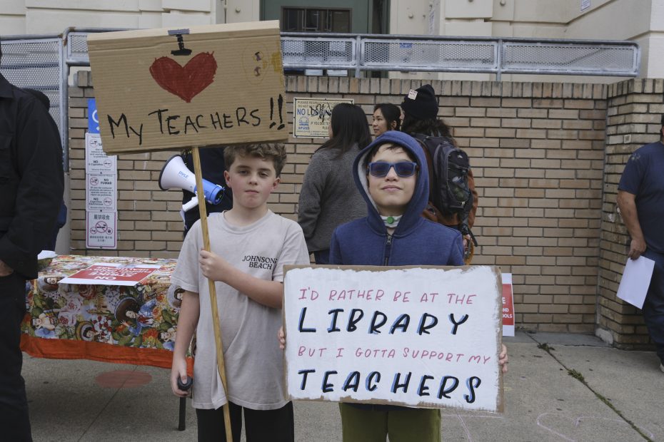 Two children stand outside holding protest signs supporting teachers; one sign says "I ♥ my teachers!!" and the other reads "I’d rather be at the library but I gotta support my teachers.
