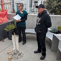 Two people stand outdoors near a small display of rocks, sticks, and fabric, holding papers. A bench, plants, and buildings are visible in the background.