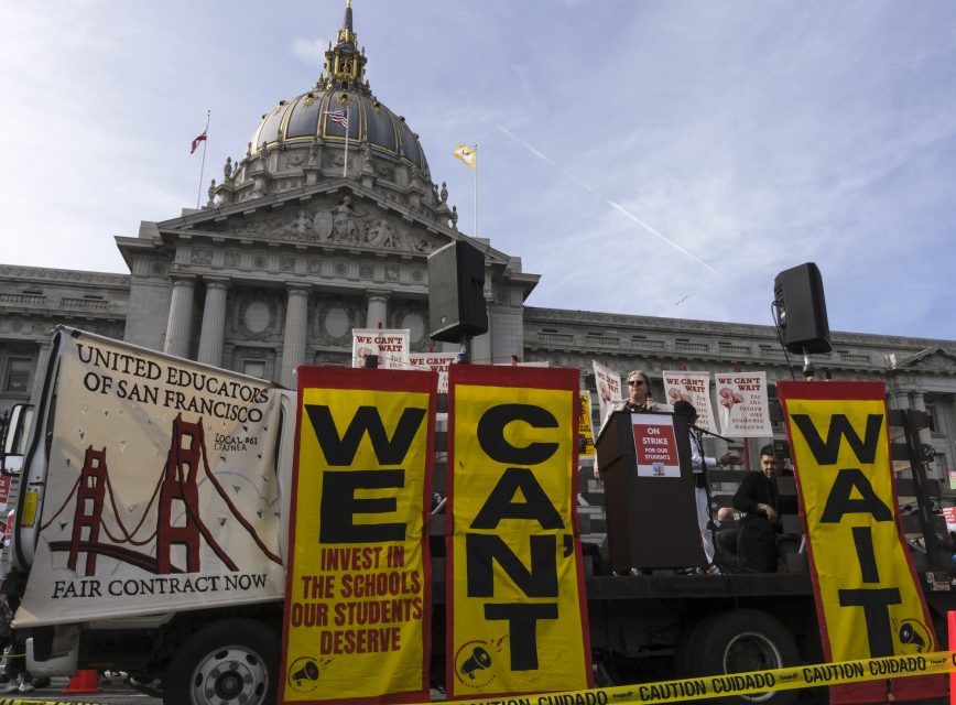 A speaker addresses a crowd at a rally in front of San Francisco City Hall. Large banners read "WE CAN'T WAIT" as teachers and supporters hold signs calling for fair contracts for educators.