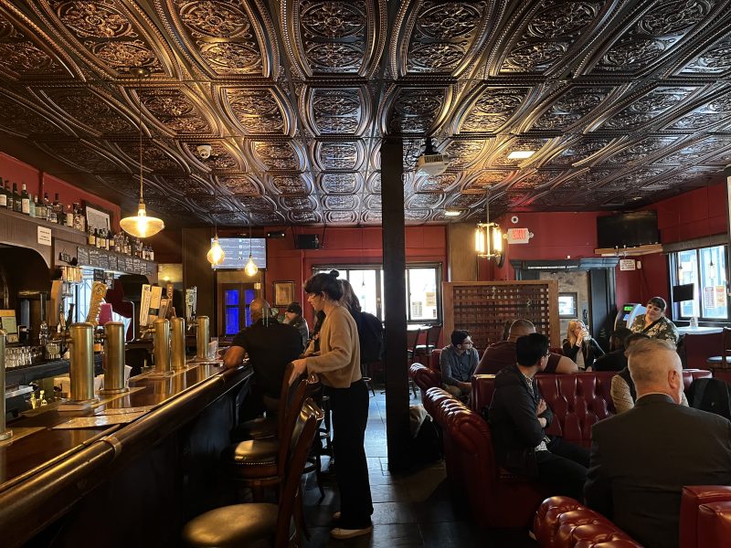 People sit and converse in a dimly lit bar with ornate tin ceiling tiles, red walls, and leather seating. A bartender serves drinks behind the counter.