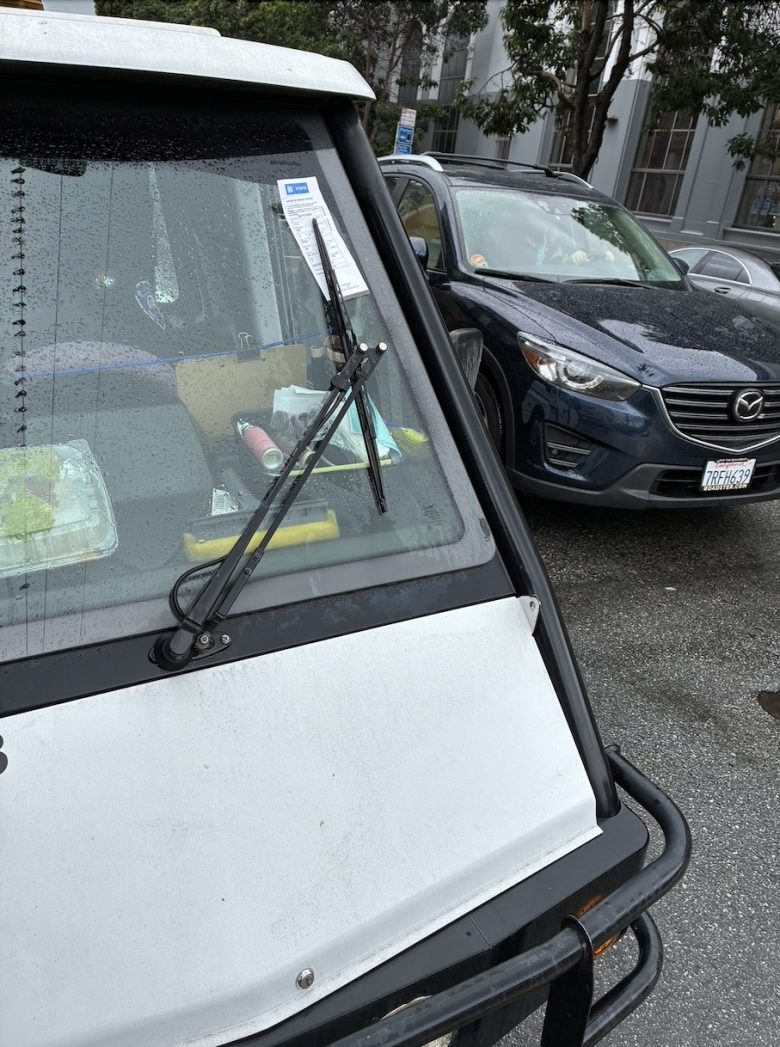 A white vehicle with a parking ticket on the windshield is parked on a wet street beside a dark gray Mazda SUV. Trees and a building are visible in the background.