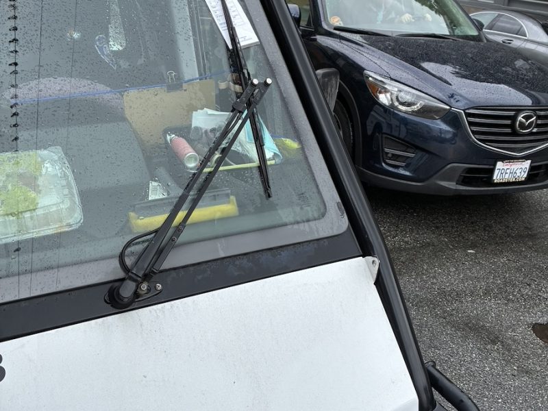 A close-up of a vehicle windshield with a single wiper, parked in a lot on a rainy day. Items are visible on the dashboard, and another car is parked nearby.