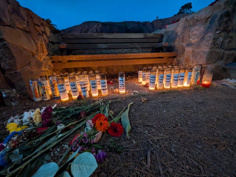 A wooden bench surrounded by lit memorial candles and scattered flowers, set against a stone wall at dusk.