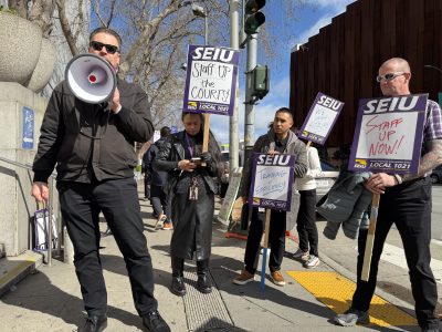 A group of people holding SEIU signs and a megaphone stand on a sidewalk during a daytime staff protest.