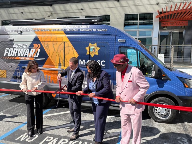 Four people cut a red ribbon in front of a van labeled "Moving Forward" during a ribbon-cutting event in a parking lot.