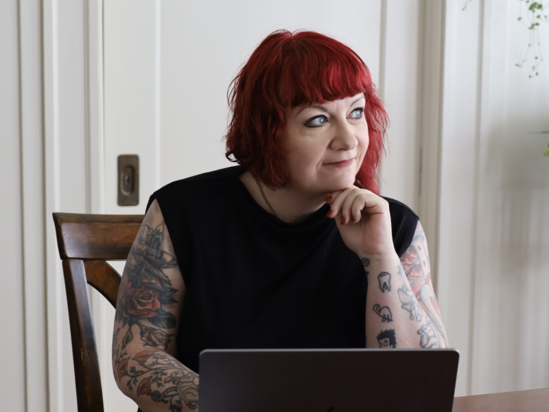 Rae Alexandra, a person with red hair and tattoos, sits at a table with a laptop, resting their chin on their hand and looking to the side.