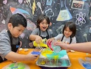 Three children pour colorful liquids into a tray while participating in a science experiment, with a chalkboard featuring scientific drawings in the background.