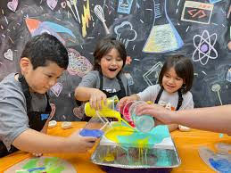 Three children pour colorful liquids into a tray during a science activity, with a chalkboard background showing drawings of lab equipment and scientific symbols.