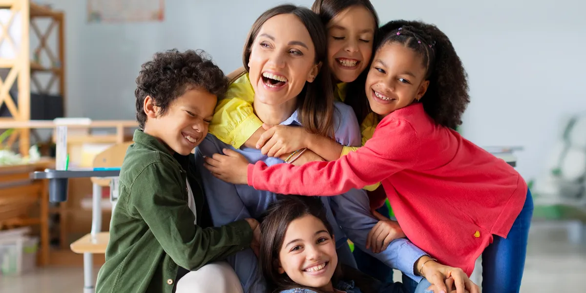 A group of four smiling children hug an adult woman while sitting together indoors, appearing happy and playful.