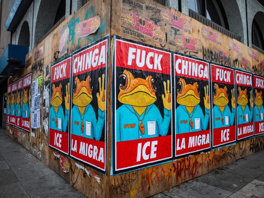 Posters with a cartoon frog in a blue shirt and gold chain display the phrases "FUCK ICE" and "CHINGA LA MIGRA" on a graffiti-covered street corner.