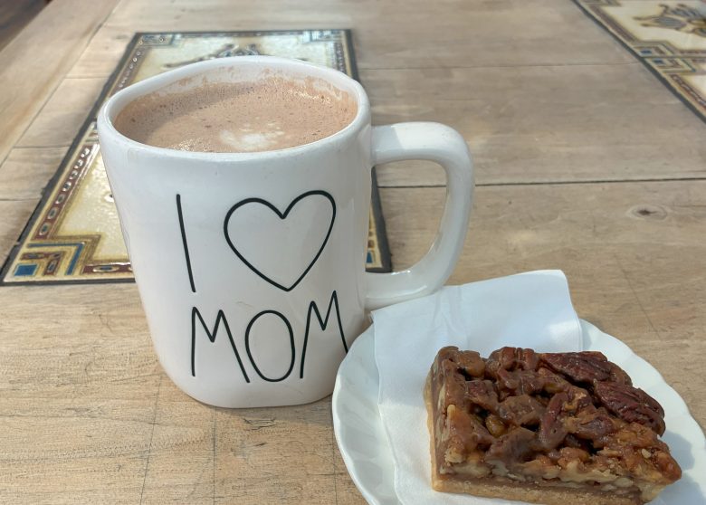 A large white mug with "I ♥ MOM" printed on it next to a slice of pecan pie on a small white plate, both placed on a wooden table.