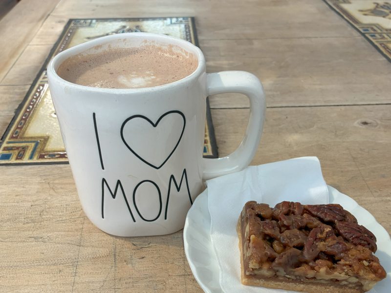 A large white mug with "I ♥ MOM" printed on it next to a slice of pecan pie on a small white plate, both placed on a wooden table.
