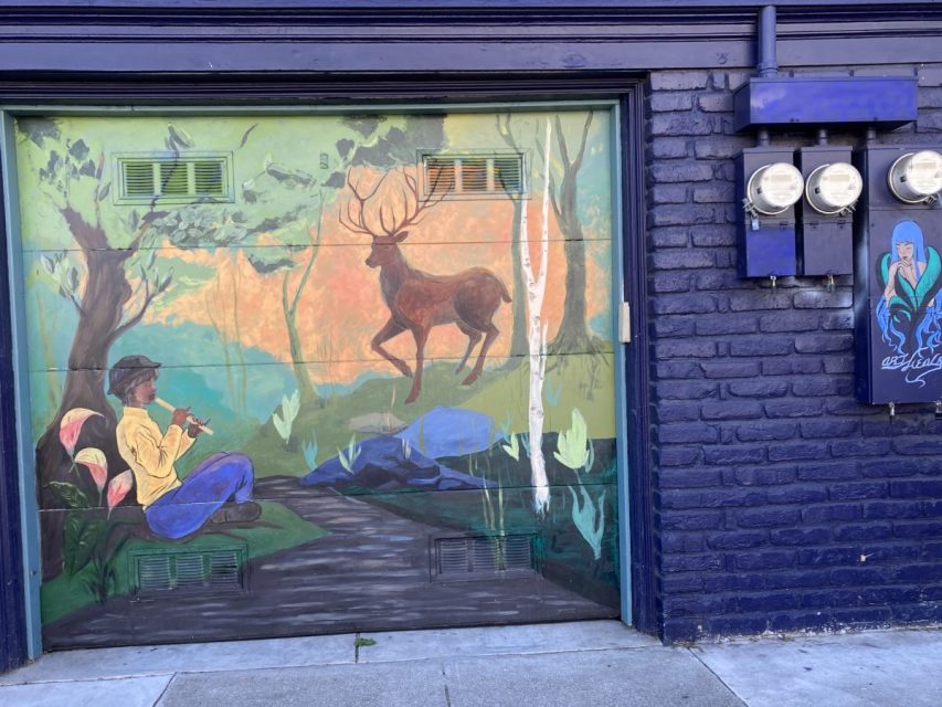 A mural on a garage door shows a person sitting and playing a flute to a deer with large antlers in a forest scene; utility meters are mounted on the adjacent brick wall.