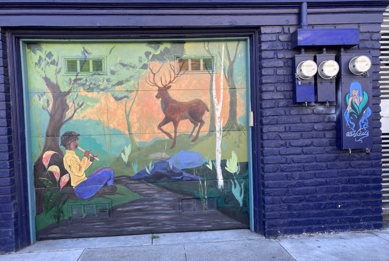 A mural on a garage door shows a person playing a flute in a forest, with a deer standing nearby. Two electric meters and a painted portrait are on the adjacent blue brick wall.