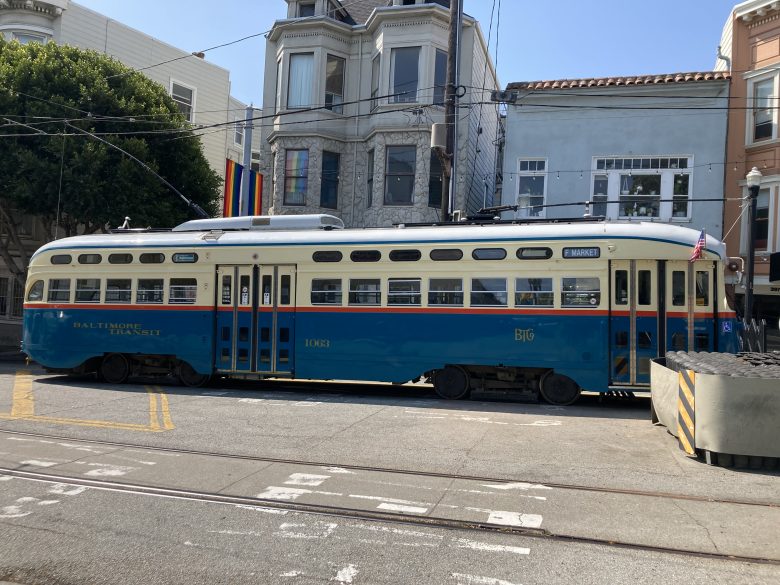 A vintage blue and cream Baltimore Transit streetcar numbered 1073 is parked on a city street, with buildings and a rainbow flag in the background.