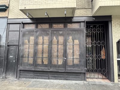 A closed storefront with windows covered by mesh and cardboard, next to a gated entrance on a city sidewalk.