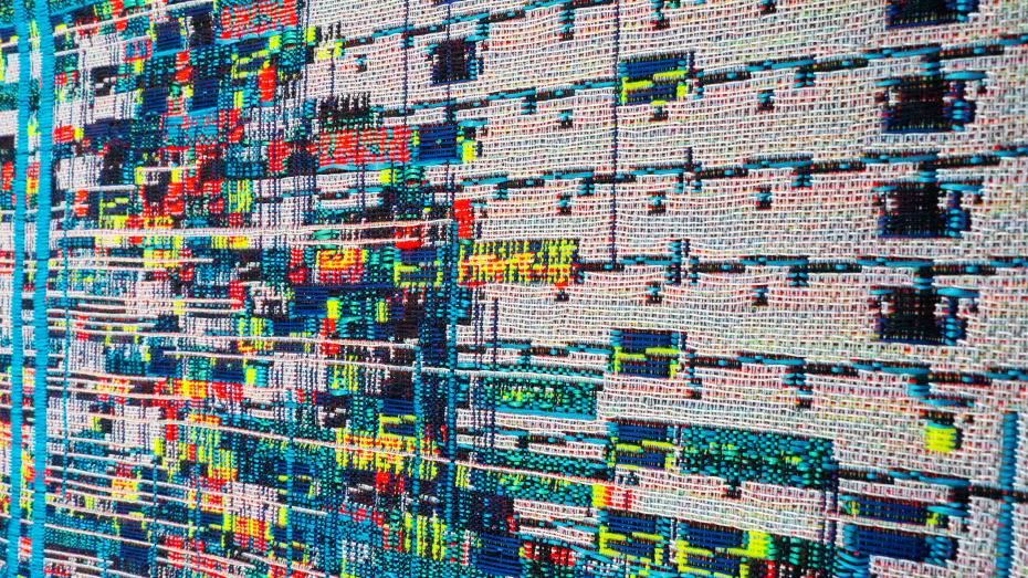 Close-up of a colorful abstract textile artwork featuring rows of multicolored threads arranged in a grid-like pattern with various geometric shapes.
