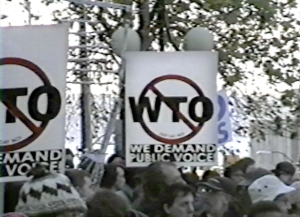 A crowd holds signs reading "WTO" crossed out and "We demand public voice" during a protest. Trees and balloons are visible in the background.