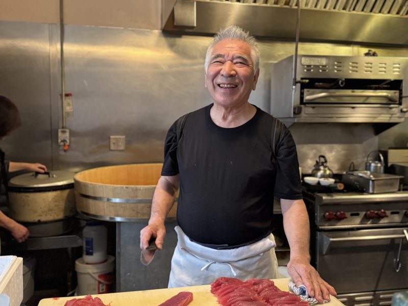 An older man in a black shirt and white apron stands in a restaurant kitchen, smiling while preparing pieces of raw fish on a counter.