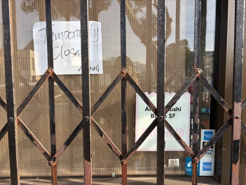 A metal security gate blocks a storefront window with a handwritten “Temporarily Closed” sign and a poster for Akari Sushi BROS SF visible behind the glass.