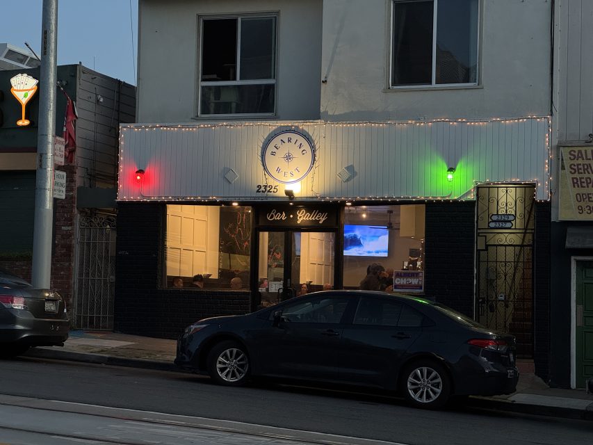 Street view of a restaurant called "Herring Nest" with red and green lights on the facade, a parked black car in front, and people seated inside.
