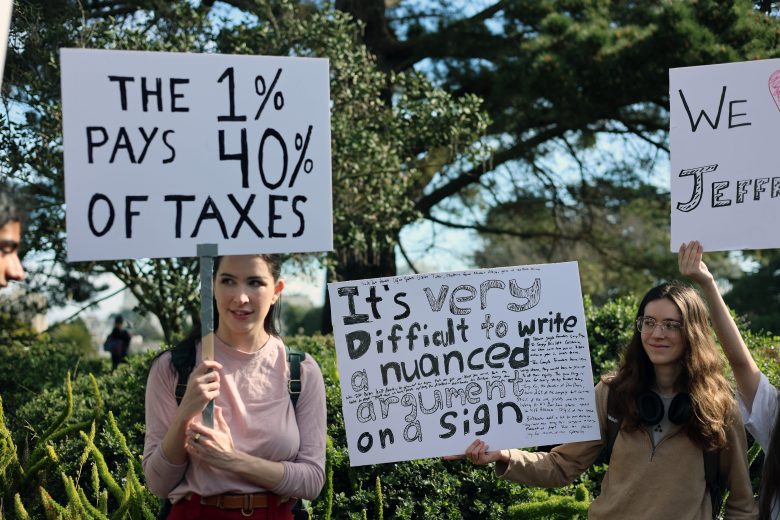 Two people at a protest hold signs, one reading "The 1% pays 40% of taxes," and the other reading "It's very difficult to write a nuanced argument on a sign.
