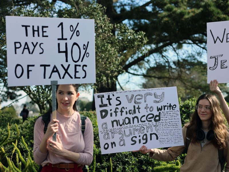 Two people at a protest hold signs, one reading "The 1% pays 40% of taxes," and the other reading "It's very difficult to write a nuanced argument on a sign.