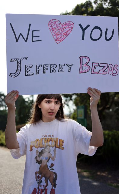 A person holds a sign that reads "We ♥ You Jeffrey Bezos" while wearing a t-shirt that says "I'm in a polycule with Bella" during the billionaire march.