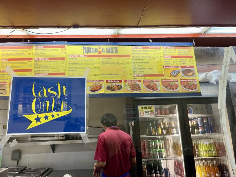 A person stands behind a counter at a restaurant with a yellow "Cash Only" sign; a large menu and refrigerated drinks are visible in the background.
