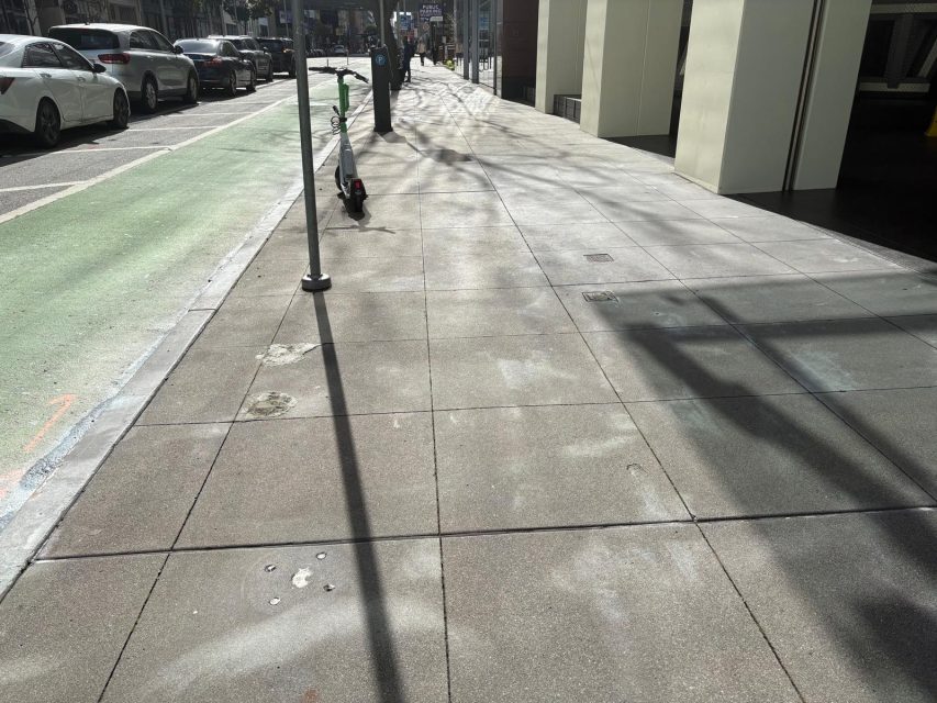 A city sidewalk with faint shadows from nearby structures, a green bike lane, parked cars, and a parked electric scooter near a pole.