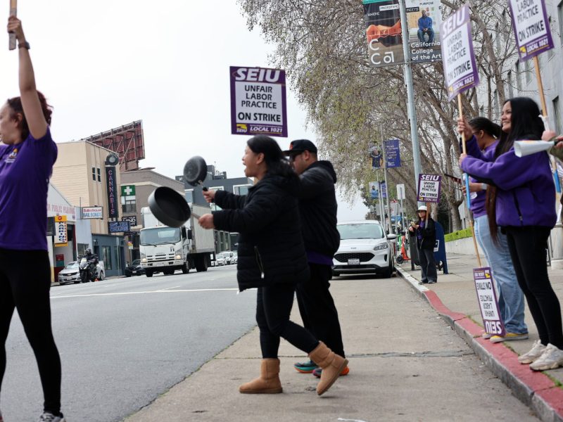 A group of people on a city sidewalk hold SEIU strike signs and bang pots, protesting unfair labor practices.
