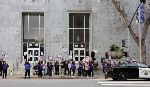 A group of people holding protest signs stands in front of a gray building; a police car is parked nearby at the curb.