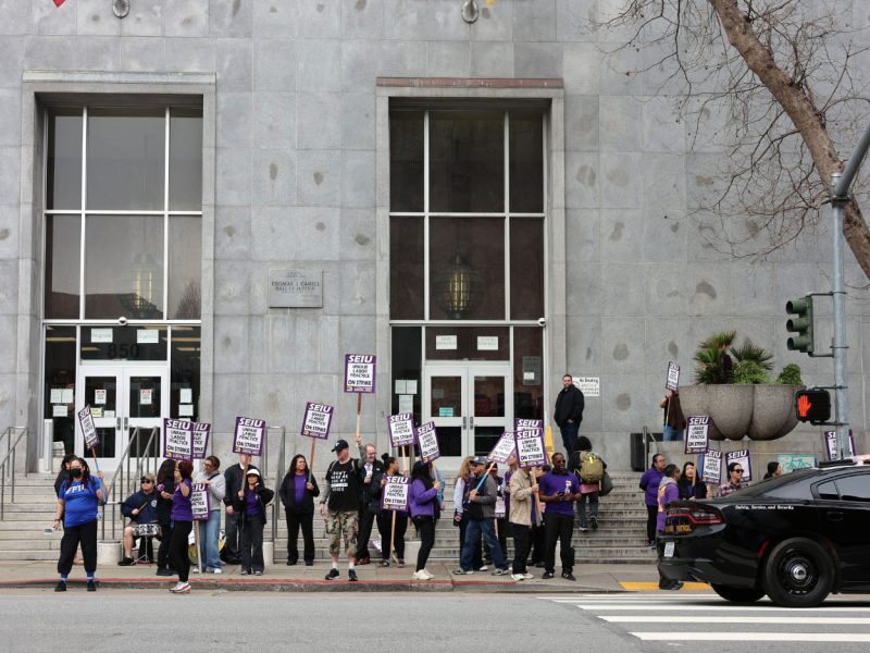A group of people holding protest signs stands in front of a gray building; a police car is parked nearby at the curb.