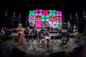 A band performs on stage with various instruments; behind them is a colorful digital backdrop featuring skull and geometric patterns.