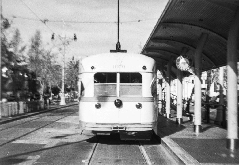 Black and white photo of a streetcar stopped at a station platform under a curved roof, with a clock visible on the right side.