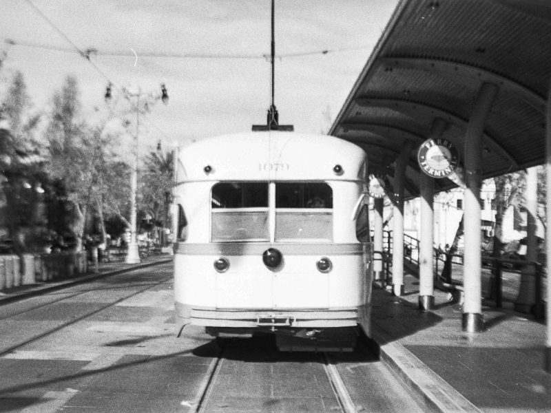 Black and white photo of a streetcar stopped at a station platform under a curved roof, with a clock visible on the right side.