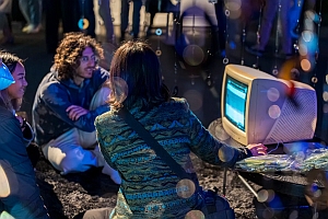 Three people sit around an old computer monitor outdoors at night, viewing its screen, with blurred lights in the foreground.