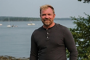 A man with blond hair and a beard stands outdoors in front of a body of water with sailboats and trees in the background.