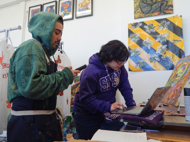 Two people work in an art studio; one uses a laptop while the other looks at a phone. Painted shirts and bee-themed artwork are visible in the background.