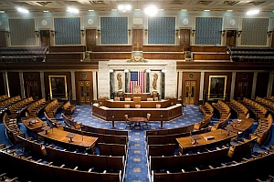 The image shows the empty chamber of the United States House of Representatives, featuring rows of desks, a podium, and an American flag at the front.